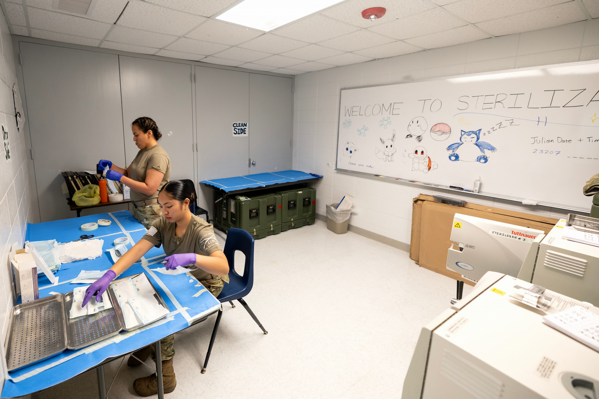 Two state workers in a work room with Sterilization procedures written on the board behind them, surrounded by supplies. 